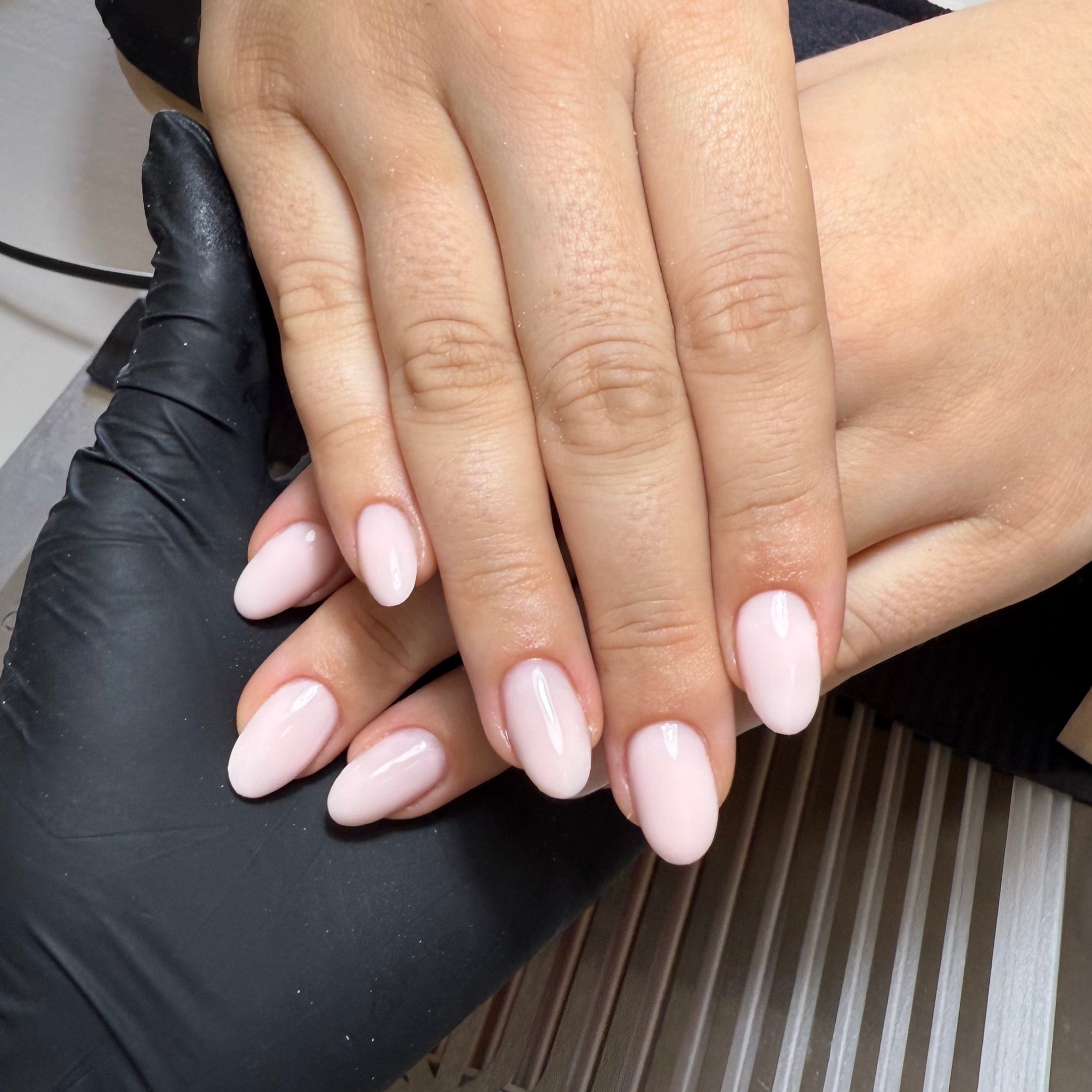 Hand with pink nail polish being worked on by a nail technician wearing black gloves.